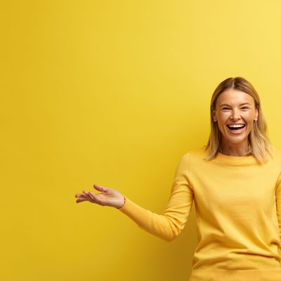 Laughing Woman Pointing Palm Away. Blonde Girl Wearing Casual Clothes Gesturing with Smile on Face at Empty Space for Your Advertisement. Indoor Studio Shot Isolated