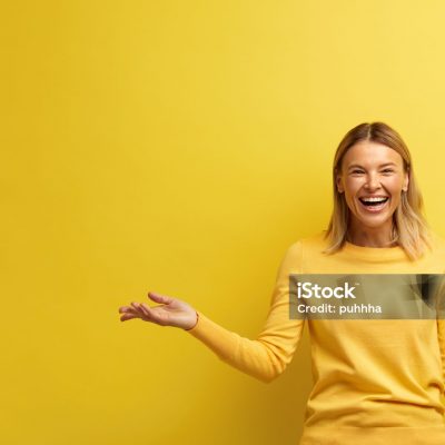 Laughing Woman Pointing Palm Away. Blonde Girl Wearing Casual Clothes Gesturing with Smile on Face at Empty Space for Your Advertisement. Indoor Studio Shot Isolated